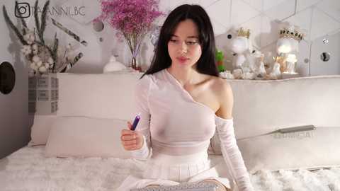 Media: Video of a slender Asian woman with long black hair, wearing a white off-shoulder top, holding a pen, sitting on a white bed with floral decor, in a softly lit bedroom.