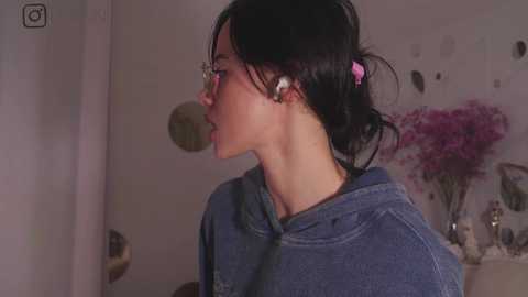 Media: Video of a young Asian woman with light skin, black hair in a pink scrunchie, wearing glasses, and a gray hoodie, in a softly lit room with a floral arrangement and abstract art on the wall.