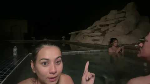 Media: Video of three women in a nighttime swimming pool, one with a puzzled expression, another holding a bottle, and a third with a raised index finger, all against a dimly lit rocky backdrop.