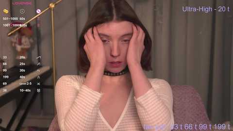 Media: Video of a young woman with shoulder-length brown hair, wearing a white striped long-sleeve top and black choker, sitting with hands on her face in a room with a purple couch and a lamp.