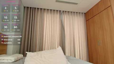 Media: Video of a modern bedroom with beige curtains covering a window, light wooden wardrobe, white bed, and white pillows.