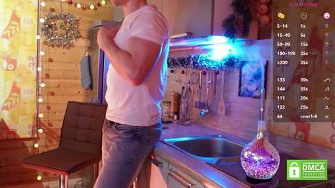 Media: Video of a muscular man in a white t-shirt and blue jeans standing in a cozy, rustic kitchen with wooden walls, hanging utensils, and a shiny sink.