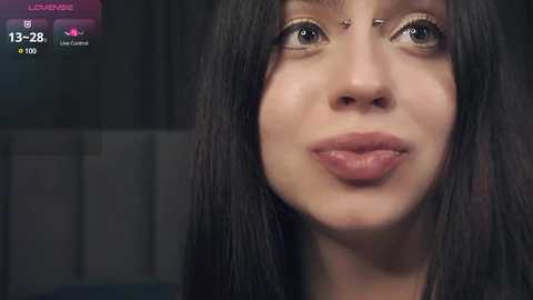 Media: Video of a young woman with light skin and long dark hair, wearing makeup and a septum piercing, smiling softly. Background is dark and blurred.