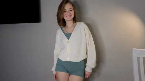 Media: A video of a young Caucasian girl with straight, light brown hair and fair skin, smiling in a casual outfit of a white cardigan over a teal dress, standing against a plain gray wall.