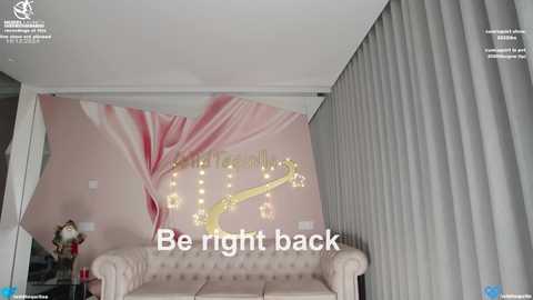 Media: Video of a modern living room with a pink and gold wall mural, a beige tufted sofa, and a \"Be right back\" message overlaid.