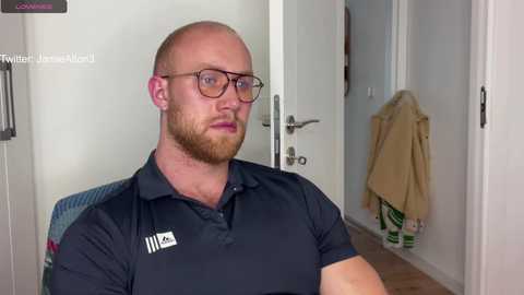 Media: Video of a bald, bearded, muscular man in glasses, wearing a dark polo shirt, seated in a hallway with beige coats hanging on a hook.