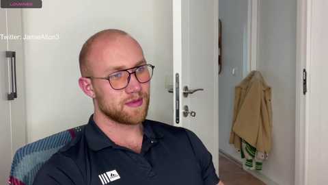 Media: Video of a bald, bearded man with glasses, wearing a dark blue collared shirt, sitting on a colorful chair. Behind him, a white door with a beige jacket hanging on the knob.