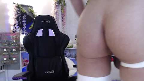 Media: Video of a nude, fair-skinned person from behind in a gaming room with a black gaming chair, a pink floral backdrop, and a monitor displaying \"10:15.\