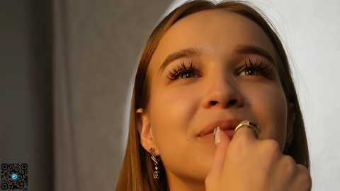 Media: A video of a young woman with fair skin, light brown hair, and brown eyes, wearing delicate earrings, holding a finger to her lips in a thoughtful pose against a neutral background.