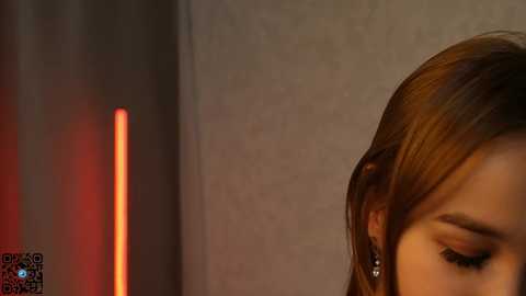 Media: Video of a woman's profile, with light brown hair, wearing earrings, against a dimly lit, textured beige wall. A vertical red light is faintly visible on the left.