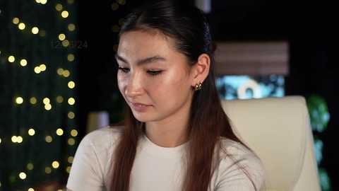 Media: Video of a young East Asian woman with long dark hair, wearing a white top and gold stud earrings, sitting indoors. Background features a blurred Christmas tree with yellow lights and a TV screen.