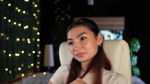 Media: Video of a young Asian woman with straight, dark hair, wearing a white t-shirt, sitting in a modern room with a Christmas tree adorned with golden lights, and a TV screen visible in the background.