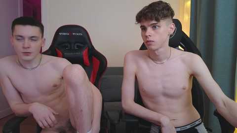 Media: Video of two young, shirtless, fair-skinned men, one with short black hair and the other with curly brown hair, sitting on a black and red gaming chair in a dimly lit room with beige walls.