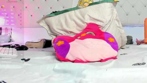 Media: Video of a pink and purple stuffed animal lying on a white bed, surrounded by various personal items, including shoes, a black hairbrush, and a white pillow.