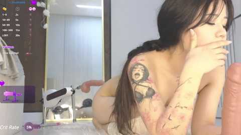 Media: Video of a nude woman with long dark hair, tattoos on her arm, and a man's erect penis in her mouth, in a modern bedroom setting with a mirror and digital clock.