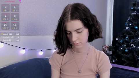 Video of a young woman with straight, shoulder-length dark hair, wearing a beige top and necklace, sitting on a bed with a blue sheet. Background features a Christmas tree and a white wall with a calendar.