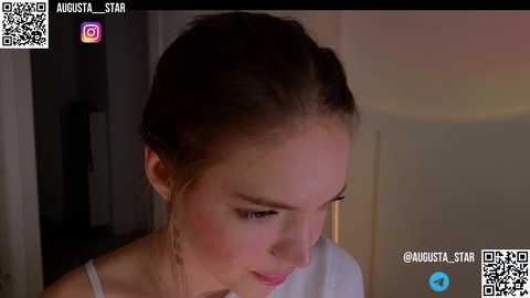 Media: Video of a young Caucasian woman with light skin, brown hair in braids, wearing a white top, looking down thoughtfully. Background is dimly lit with a blurred wall.