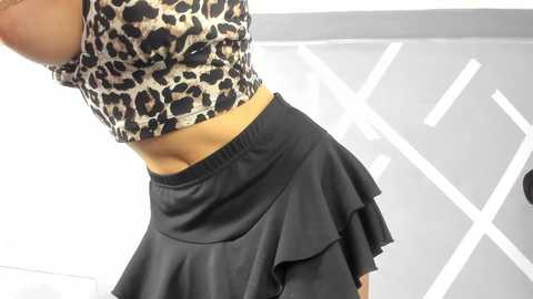 Media: Video of a woman lifting her leopard-print crop top, revealing her midriff. She wears a black, ruffled skirt. Background shows a grey wall with white geometric patterns.