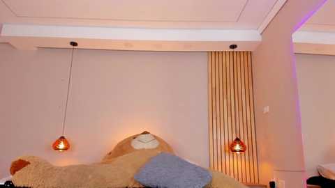 Media: Video of a cozy bedroom with a plush, beige bear-shaped bed, adorned with two brown and blue pillows. Two orange pendant lights hang from the ceiling, casting a warm glow, and a vertical wooden panel adds texture to the plain walls.