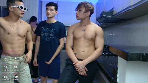 Media: Video of three shirtless young men in a modern kitchen, one with sunglasses, one in a black graphic tee, and one in black pants, under blue lighting.