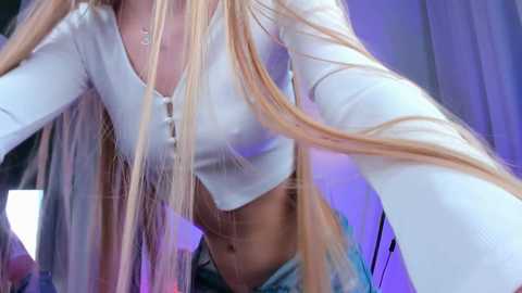 Media: Video of a blonde woman with long hair, wearing a white crop top and blue jeans, captured in a dimly lit room with purple lighting.