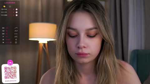 Media: Video of a young woman with long, straight blonde hair, wearing minimal makeup, sitting indoors. Background features a modern lamp and soft, neutral-toned decor.