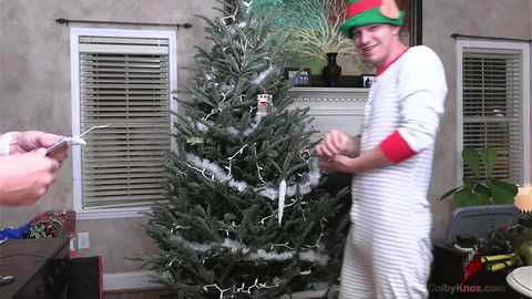 Media: Video of a man in a white and red striped onesie, holding a gift, standing beside a decorated Christmas tree in a living room with beige walls and blinds.