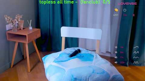 Media: Video of a cozy bedroom with a white chair, a blue and white blanket, wooden side table with plant, teal curtains, wooden floor, and a black vibrator on the blanket.