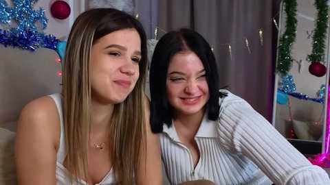 Media: Video of two young women smiling, one with long blonde hair in a white tank top, the other with black hair in a white sweater, in a festive, decorated room.