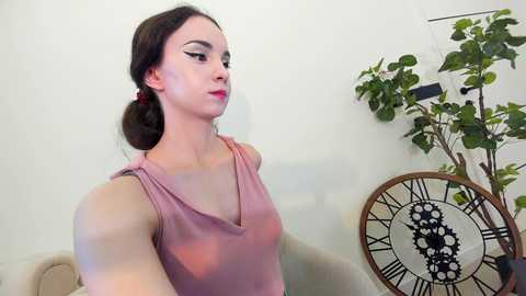 Media: Video of a fair-skinned woman with brown hair in a ponytail, wearing a pink sleeveless top, standing next to a decorative clock and green plant against a plain white wall.