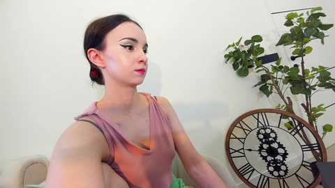 Media: Video of a fair-skinned woman with dark hair, wearing a pink tie-dye top, posing in a minimalist room with a green plant and a clock.