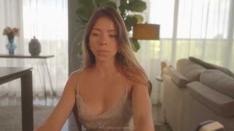 Media: Video of a young Asian woman with long, straight brown hair, wearing a beige lace camisole, sitting in a modern living room with large windows, potted plants, and a gray sofa.