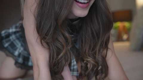 Media: Video of a young woman with long, wavy brown hair, wearing a black and white plaid dress, smiling widely with visible teeth, leaning forward in a cozy, dimly lit room.