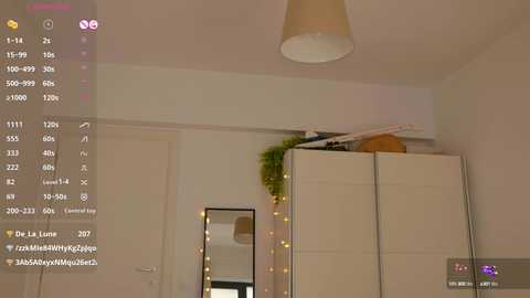 Media: A video captures a minimalist bedroom with beige walls, a white wardrobe, and a small, rectangular mirror. A hanging plant adds greenery. A ceiling lamp casts soft light, and a smartphone display is visible in the top left corner.