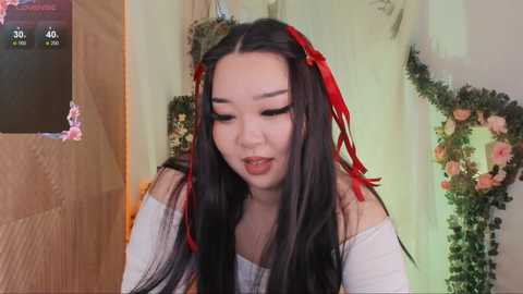 Media: A video of an East Asian woman with long, black hair adorned with red ribbons, wearing a white off-shoulder top, in a softly lit room with floral decorations.