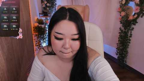 Media: Video of a plus-size East Asian woman with long black hair, wearing white off-shoulder top, sitting in a chair with a serene expression. Background features floral decorations and a digital screen displaying a clock.