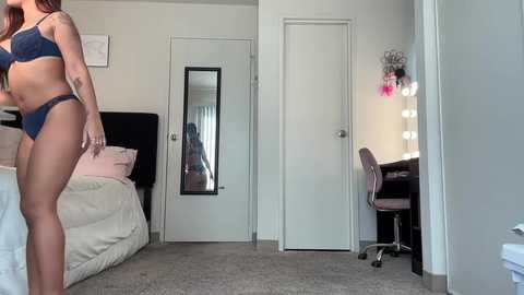 Media: Video of a modern bedroom with a woman in blue lingerie, standing near a bed, mirror, and vanity table with pink flowers.