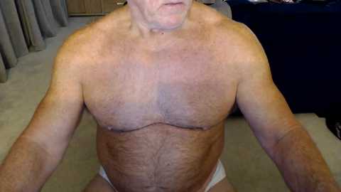 Media: Video of a middle-aged, light-skinned, shirtless man with a hairy chest and abdomen, wearing white briefs, sitting on a beige carpet in a dimly lit room with wooden furniture and dark curtains.