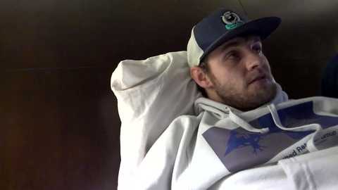 Media: Video of a Caucasian man with a beard, wearing a blue cap, white hoodie, and blue shirt, reclining in a car, looking introspective.