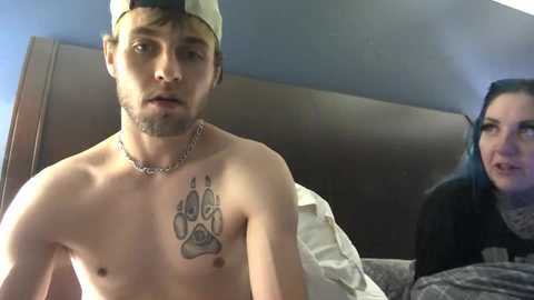 Media: Video of a shirtless, pale young man with a tattoo of a paw on his chest, wearing a backward baseball cap, sitting on a bed in a dimly lit room.