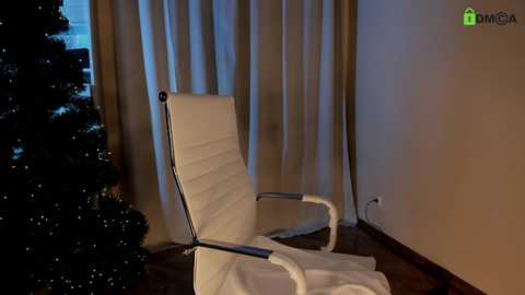 A video of a modern white recliner chair with a white blanket draped over it, positioned in a dimly lit room with beige curtains and a dark, glittery Christmas tree partially visible on the left.