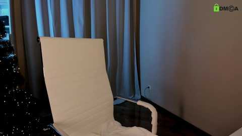 Media: A video of a modern, minimalist hotel room with a white, cushioned lounge chair against dark, shiny curtains; soft, warm lighting creates a cozy ambiance.