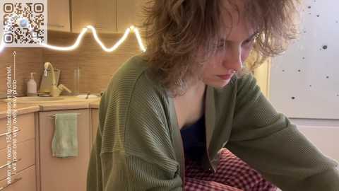 Media: Video of a curly-haired woman in a green cardigan, sitting on a red and white checkered blanket in a cozy kitchen with beige cabinets and a glowing neon light.