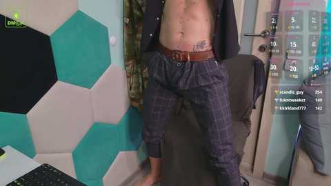 Media: Video of a shirtless man with a tattoo on his lower abdomen, wearing plaid pajama pants, standing in a modern room with hexagonal teal and white wall panels and a digital clock displaying 15:40.