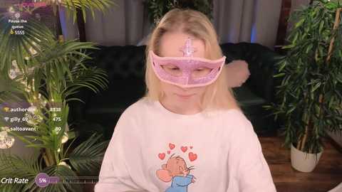 Media: Video of a blonde woman in a pink masquerade mask, smiling, wearing a \"Tangled\" shirt, surrounded by lush green plants.