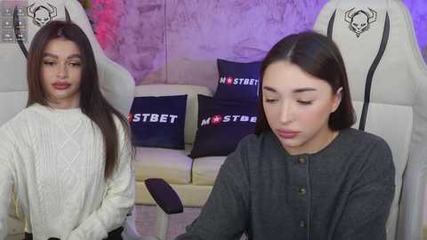 Media: Video of two young women with long hair, one in white sweater, the other in gray, seated in gaming chairs with black pillows.