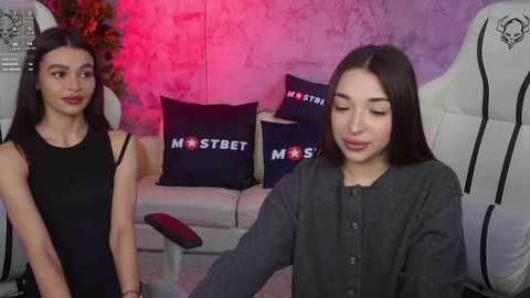 Media: Video of two women in a modern living room. One wears a black sleeveless dress, the other a grey cardigan. Dark pillows with \"MST3K\" logos on a white couch.