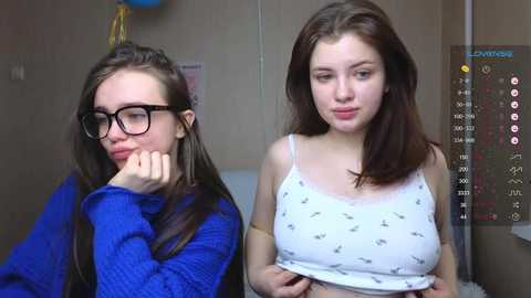 Media: Video of two teenage girls with long brown hair, one in glasses, blue sweater, and the other in a white crop top with butterflies, sitting side by side in a classroom.