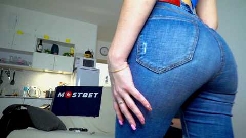 Media: Video of a woman's rear in tight blue jeans, standing in a modern kitchen with white cabinets, a fridge, and a \"MOSTBET\" laptop on a counter.