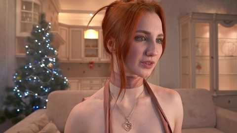 Media: Video of a fair-skinned, red-haired woman in a white dress, with a necklace, in a cozy, softly-lit living room featuring a decorated Christmas tree and glass cabinet.